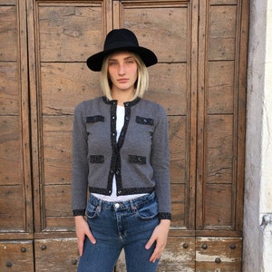 Puede incluir: Un cárdigan gris con ribetes negros y detalles brillantes. El cárdigan tiene un cuello redondo y cierre de botones. La modelo lleva un sombrero fedora negro y unos vaqueros azules.