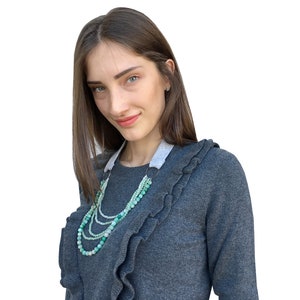 Puede incluir: Una mujer con un su&eacute;ter gris con volantes y un collar de cuentas verdes y blancas de varias capas.