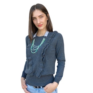 Puede incluir: Una mujer con un su&eacute;ter gris con cuello de volantes y un collar de cuentas verdes y blancas en capas. Lleva pantalones vaqueros azules y tiene las manos en los bolsillos.