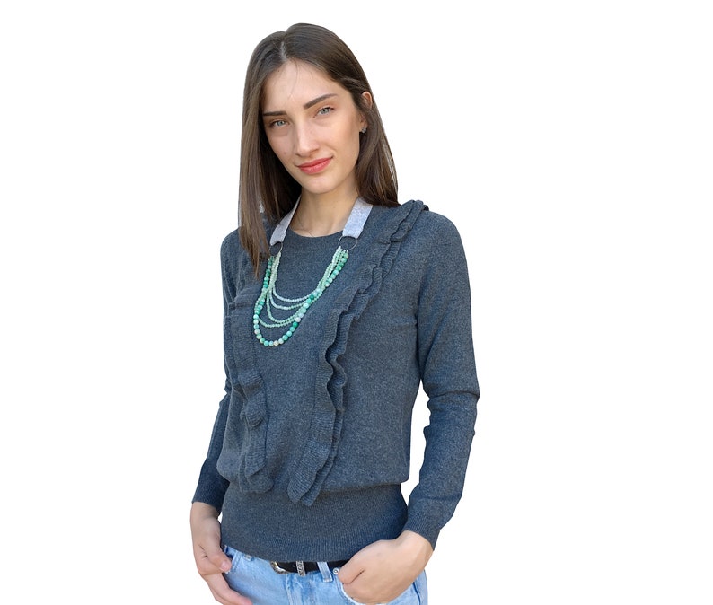 Puede incluir: Una mujer con un su&eacute;ter gris con cuello de volantes y un collar de cuentas verdes y blancas en capas. Lleva pantalones vaqueros azules y tiene las manos en los bolsillos.
