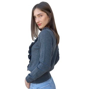 Puede incluir: Una mujer con un su&eacute;ter gris de manga larga con cuello de volantes y jeans azules. El su&eacute;ter tiene una silueta ajustada y mangas largas.