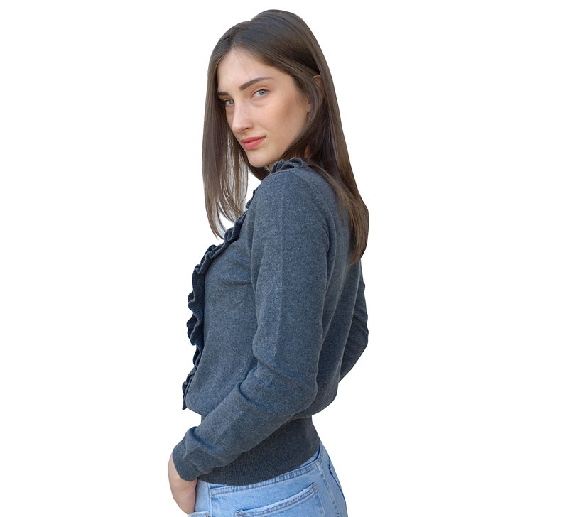 Puede incluir: Una mujer con un su&eacute;ter gris de manga larga con cuello de volantes y jeans azules. El su&eacute;ter tiene una silueta ajustada y mangas largas.