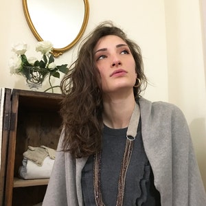 Puede incluir: Una mujer lleva un c&aacute;rdigan gris sobre un jersey gris con volantes y un collar de cuentas de m&uacute;ltiples hilos. El collar tiene un anillo de plata y una correa de cuero marr&oacute;n.