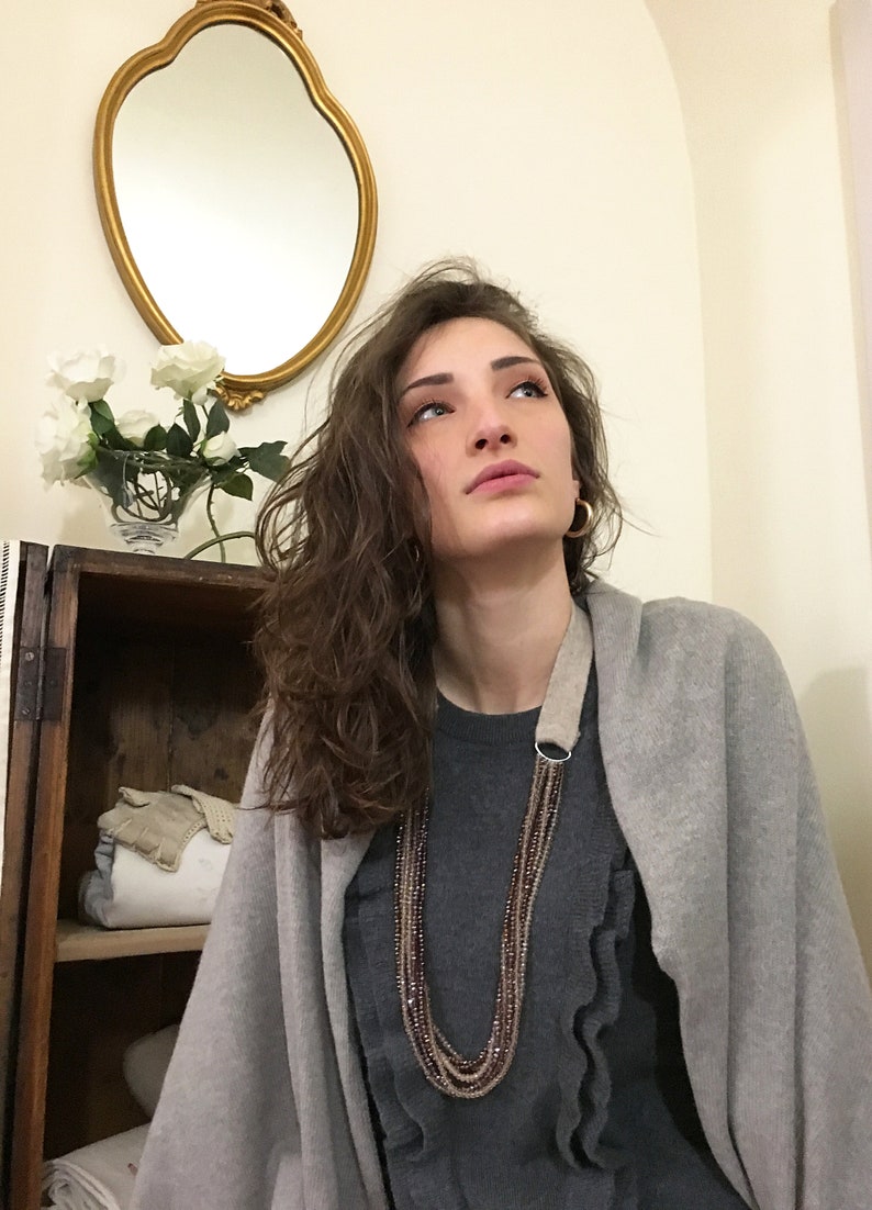 Puede incluir: Una mujer lleva un c&aacute;rdigan gris sobre un jersey gris con volantes y un collar de cuentas de m&uacute;ltiples hilos. El collar tiene un anillo de plata y una correa de cuero marr&oacute;n.