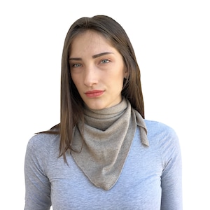 Peut inclure: Un foulard triangulaire marron clair est porté autour du cou. Le foulard est fait d'une matière douce et a un design simple. La personne sur l'image porte un haut à manches longues gris.