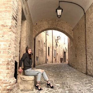 Puede incluir: Una mujer con un su&eacute;ter gris, jeans rotos y sandalias de tac&oacute;n negro se sienta en un escal&oacute;n de piedra en un callej&oacute;n estrecho y abovedado. El callej&oacute;n est&aacute; bordeado de edificios de piedra y tiene un suelo de adoquines.