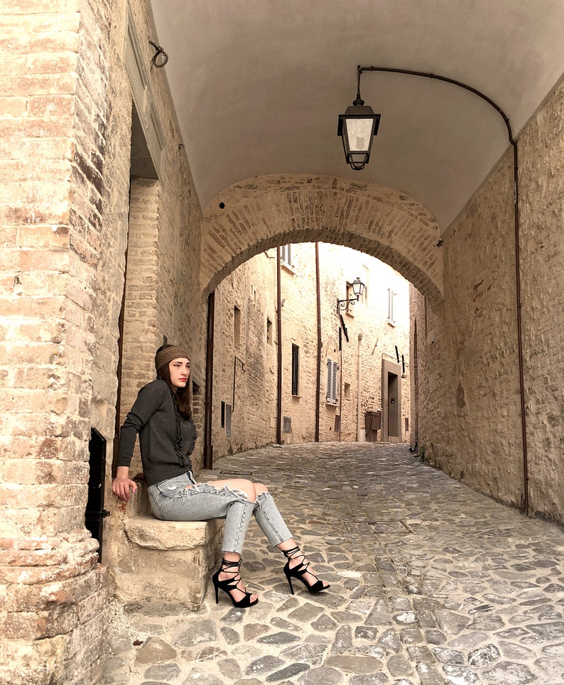 Puede incluir: Una mujer con un su&eacute;ter gris, jeans rotos y sandalias de tac&oacute;n negro se sienta en un escal&oacute;n de piedra en un callej&oacute;n estrecho y abovedado. El callej&oacute;n est&aacute; bordeado de edificios de piedra y tiene un suelo de adoquines.