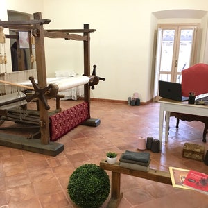 Puede incluir: Una habitaci&oacute;n con un gran telar de madera, una silla roja, un escritorio blanco con un port&aacute;til y una variedad de hilo y tela. La habitaci&oacute;n tiene un suelo de baldosas de terracota y una gran ventana.