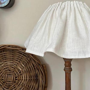 May include: A white linen lampshade on a dark wooden lamp base. A round, woven wicker tray with handles is in the foreground. A dark metal object hangs on the wall in the background. The scene has a rustic, farmhouse aesthetic.