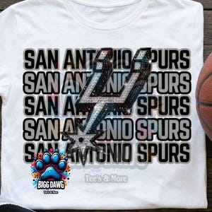 May include: White t-shirt featuring the text "SAN ANTONIO SPURS" in a stacked design with a silver and black Spurs logo. The shirt also includes a colorful paw print graphic and the text "BIGG DAWG Tee's & More".
