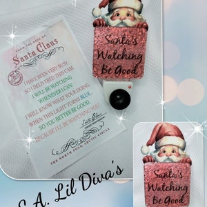 May include: A Christmas-themed night light featuring Santa Claus holding a sign that reads "Santa's Watching Be Good." The night light has a red glittery sign and a white base with a black lens. A letter from Santa is also included.