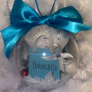 Puede incluir: Un adorno de Navidad de vidrio transparente con un lazo de cinta azul y una bolsa de regalo Tiffany & Co. en miniatura en su interior. El adorno está lleno de nieve artificial blanca.