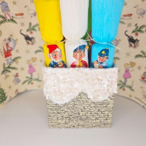 Puede incluir: Un recipiente decorativo con un exterior texturizado similar a la piedra, rematado con un ribete blanco y brillante. Contiene tubos de papel de colores y pequeñas figuras ilustradas de niños con atuendo festivo. El fondo presenta un patrón vintage con temática navideña.