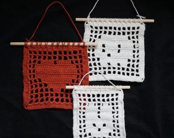 May include: Three square crochet wall hangings suspended from wooden dowels with yarn loops. One is rust-colored with a heart pattern, while two are white, featuring a ghost design and a smaller ghost motif, all with openwork grid borders.