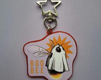 May include: A Halloween-themed keychain featuring a cartoon bee dressed as a ghost. The keychain has a star-shaped clasp and a red border. The bee is on a light purple background with the words "BOO BEE".