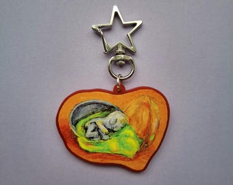 May include: A heart-shaped keychain with a painted image of a fetus inside a womb. The keychain has a star-shaped clasp and a red border. The fetus is depicted in white and gray, resting on a green blanket.