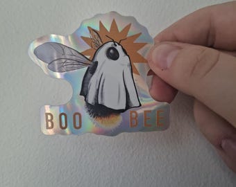 May include: Holographic sticker featuring a cartoon bee dressed as a ghost. The bee has black and white markings, wings, and a stinger. The sticker has the words "BOO BEE" in a playful font, with a starburst design behind the bee.