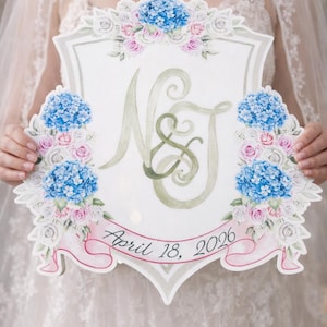May include: A white wedding sign in a shield shape, decorated with blue hydrangeas, pink and white roses, and green leaves. The sign has the initials "N & J" and the date "April 18, 2026" written on it.