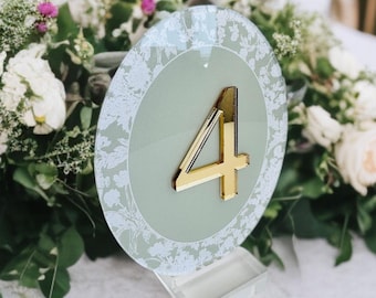 May include: A circular table number sign featuring a sage green center with a white floral border and a reflective gold acrylic number four attached to the front, supported by a clear acrylic stand on a white tablecloth.