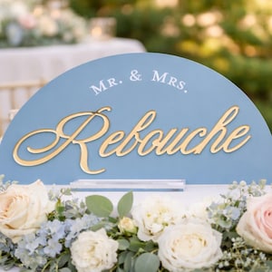 May include: A pale blue wedding sign with gold script reading "Mr. & Mrs. Rebouche." The sign is surrounded by a floral display of white and pink roses, and blue hydrangeas. Two glass candle holders with candles are visible.