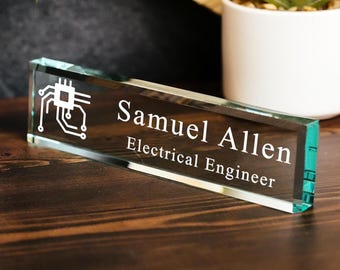 May include: A rectangular glass desk nameplate featuring a white etched circuit board icon on the left side, followed by the text Samuel Allen and the title Electrical Engineer in a clean, white, sans-serif font on the front surface.