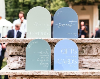 May include: Four arched acrylic signs in shades of sage green and dusty blue. The signs feature white printed text including "Please sign GUESTBOOK," "LOVE IS sweet TAKE A TREAT," "OPEN BAR THE drinks ARE ON US BUT THE hangover IS ON YOU," and "GIFTS and CARDS."