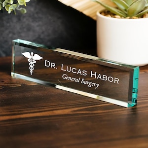 Puede incluir: Placa de escritorio rectangular de cristal transparente con el texto "DR. LUCAS HABOR General Surgery" en blanco. Un símbolo de caduceo blanco está a la izquierda. El borde de la placa tiene un tinte verde.