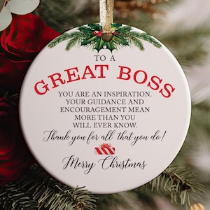 Może przedstawiać: Okrągła biała ceramiczna ozdoba ze złotą wstążką i grafiką ostrokrzewu na górze. Tekst brzmi: TO A GREAT BOSS, YOU ARE AN INSPIRATION. YOUR GUIDANCE AND ENCOURAGEMENT MEAN MORE THAN YOU WILL EVER KNOW. Thank you for all that you do! Merry Christmas, z małą ikoną czerwono-białego prezentu w paski.
