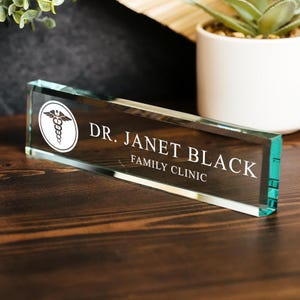 May include: A rectangular glass desk nameplate with the text "DR. JANET BLACK FAMILY CLINIC" in white. The nameplate has a medical symbol in a white circle on the left side. The nameplate is on a wooden surface.