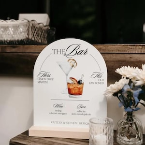 May include: An arched white bar menu sign with a wooden base featuring illustrations of a martini and an old fashioned. The text reads THE BAR, HERS LEMON DROP MARTINI, HIS OLD FASHIONED, WINE, BEER, and KAITLYN & STEVEN | 05.30.26.