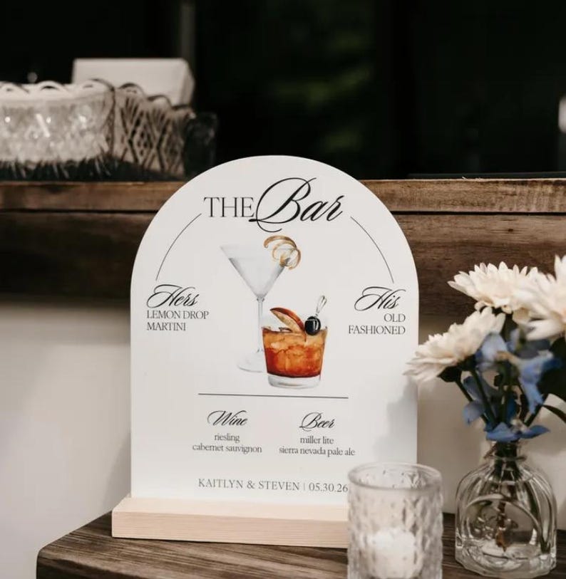 May include: An arched white bar menu sign with a wooden base featuring illustrations of a martini and an old fashioned. The text reads THE BAR, HERS LEMON DROP MARTINI, HIS OLD FASHIONED, WINE, BEER, and KAITLYN & STEVEN | 05.30.26.
