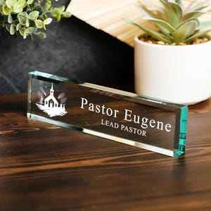 May include: A rectangular glass nameplate with the text "Pastor Eugene, LEAD PASTOR" in white. The nameplate features a church graphic and is displayed on a wooden surface.
