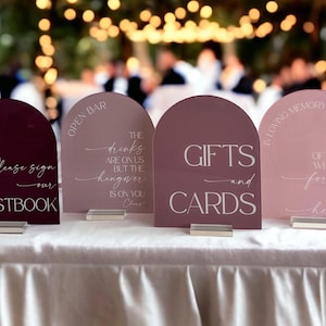 May include: Four arched acrylic signs in shades of burgundy, mauve, and blush pink. The signs feature white text reading Please sign our GUESTBOOK, OPEN BAR THE drinks ARE ON US BUT THE hangover IS ON YOU, GIFTS and CARDS, and IN LOVING MEMORY OF THOSE WHO ARE forever IN OUR hearts.