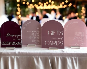 May include: Four arched acrylic signs in shades of burgundy, mauve, and blush pink. The signs feature white text reading Please sign our GUESTBOOK, OPEN BAR THE drinks ARE ON US BUT THE hangover IS ON YOU, GIFTS and CARDS, and IN LOVING MEMORY OF THOSE WHO ARE forever IN OUR hearts.