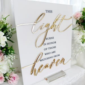 May include: A white rectangular sign featuring the text "THIS light BURNS IN HONOR OF THOSE WHO ARE WATCHING FROM heaven". The words "light" and "heaven" are rendered in a raised, reflective gold script, while the remaining text is in black.
