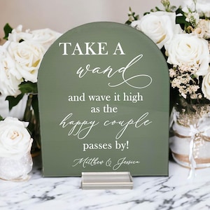 May include: An arched olive green sign with white text that reads: TAKE A wand and wave it high as the happy couple passes by! Matthew & Jessica. The sign is supported by a clear rectangular base and features a mix of serif and script fonts.