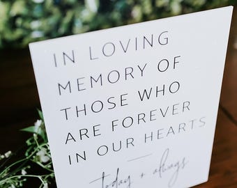 May include: A white rectangular sign with black text that reads IN LOVING MEMORY OF THOSE WHO ARE FOREVER IN OUR HEARTS and today + always in a script font. The sign is supported by a small rectangular base and is positioned near white flowers.