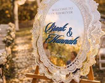 May include: An oval mirror wedding sign with a white laser-cut lace-style frame. The text reads WELCOME TO OUR FOREVER, Brook & Hannah, and EST. 02.23.2022. The sign is mounted on a wooden tripod easel, reflecting the surrounding outdoor scenery.