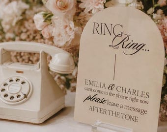 May include: A cream-colored rotary telephone next to an arched acrylic sign with black text that reads: RING Ring... EMILIA & CHARLES can't come to the phone right now please leave a message AFTER THE TONE. The sign sits on a clear base.