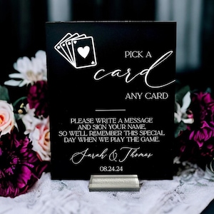 May include: A rectangular black sign with white text and a playing card graphic. The text reads: PICK A card ANY CARD, PLEASE WRITE A MESSAGE AND SIGN YOUR NAME, SO WE'LL REMEMBER THIS SPECIAL DAY WHEN WE PLAY THE GAME. Sarah & Thomas 08.24.24.