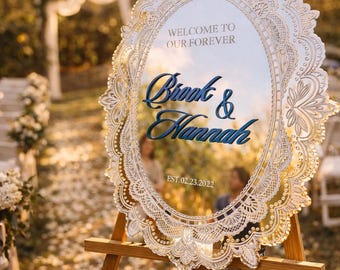May include: An oval mirror wedding sign with an intricate white laser-cut lace-style border. The text reads WELCOME TO OUR FOREVER, Brook & Hannah, and EST. 23.02.2022. The sign is mounted on a wooden tripod easel in an outdoor setting.