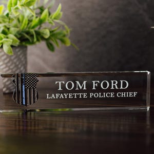 Pode incluir: Uma placa de mesa de vidro transparente com o nome "TOM FORD" e o título "LAFAYETTE POLICE CHIEF" em letras brancas. Um distintivo da polícia com uma linha azul e um design de bandeira americana está à esquerda.