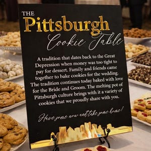 May include: A black rectangular sign with gold lettering and a gold skyline base. The text reads: The Pittsburgh Cookie Table, followed by a paragraph explaining the tradition, and the phrase: Have some now and take some home!