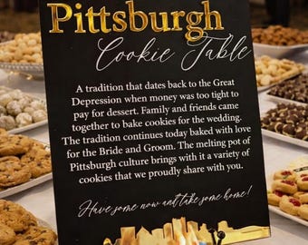 May include: A black rectangular sign with gold lettering and a gold skyline base. The text reads: The Pittsburgh Cookie Table, followed by a paragraph explaining the tradition, and the phrase: Have some now and take some home!