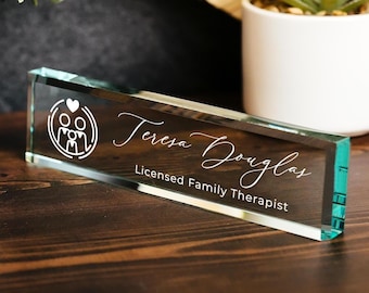 Puede incluir: Una placa de escritorio rectangular de vidrio con el texto grabado en blanco Teresa Douglas y Licensed Family Therapist, acompañada de un icono gráfico blanco de tres figuras estilizadas bajo un símbolo de corazón.