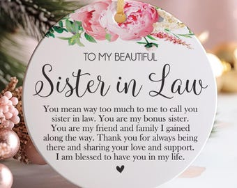 Könnte beinhalten: Ein rundes, weißes Keramikornament mit goldenem Band und einem Blumenmuster aus rosa Pfingstrosen und grünen Blättern. Der Text lautet: TO MY BEAUTIFUL Sister in Law, gefolgt von einer herzlichen Botschaft über Freundschaft und einem kleinen schwarzen Herz.
