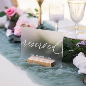 May include: A rectangular frosted acrylic sign held by a light wood base. The sign features the white cursive text "reserved" above the white printed text "FOR THE NABER FAMILY". The sign is positioned upright on a surface with a soft texture.