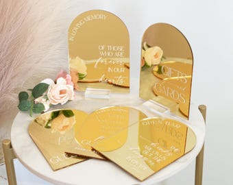May include: A set of gold arched acrylic signs with white lettering. Two signs stand upright on clear bases, while three lie flat. Text includes IN LOVING MEMORY OF THOSE WHO ARE FOREVER IN OUR HEARTS, GIFTS AND CARDS, OPEN BAR, and GUESTBOOK.