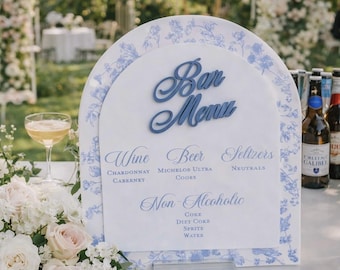 Puede incluir: Un letrero de menú de bar en forma de arco con fondo blanco y borde floral azul. El letrero muestra el texto Bar Menu, Wine, Beer, Seltzers y Non-Alcoholic, seguido de una lista de opciones de bebidas que incluyen Chardonnay, Cabernet, Michelob Ultra, Coors, Neutrals, Coke, Diet Coke, Sprite y Water.