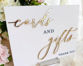 May include: A white rectangular sign featuring the gold-colored 3D script text cards and gifts, with the word AND in dark blue capital letters and the text THANK YOU in dark blue capital letters, supported by a clear acrylic base stand.